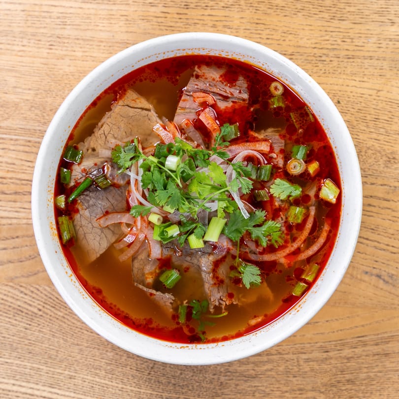 A bowl of spicy Vietnamese pho with beef, noodles, cilantro, and green onions on a wooden table.
