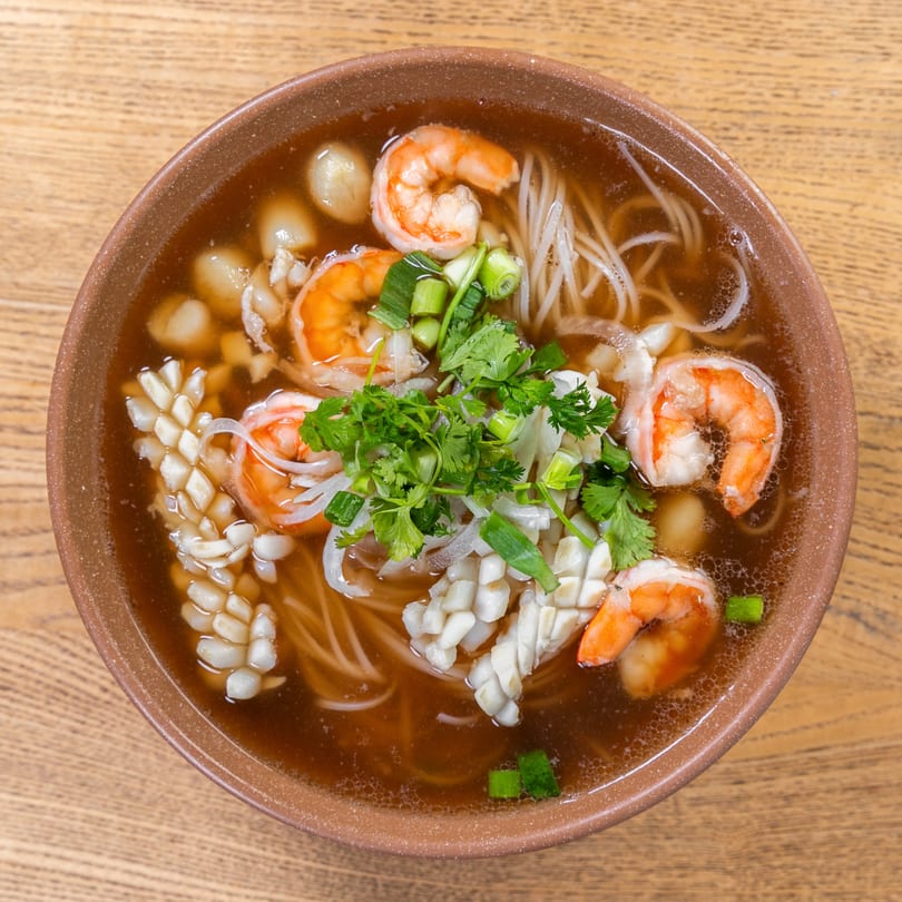 Vietnamese seafood noodle soup with shrimp, squid, and herbs in a brown bowl.